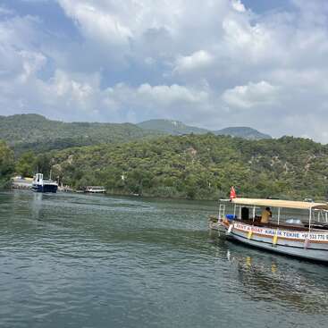 Una tranquila escena fluvial con barcas, montañas verdes al fondo y un cielo nublado. Hay una barca de alquiler con bandera turca amarrada. Ambiente tranquilo.