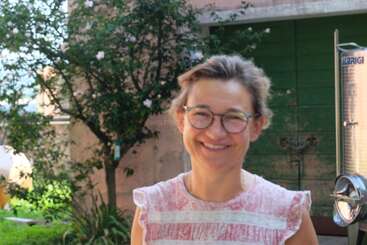 Eine lächelnde Frau mit Brille und einer rosa Bluse steht draußen. Hinter ihr sind eine grüne Tür, ein blühender Baum, Pflanzen und ein glänzender Metalltank zu sehen.