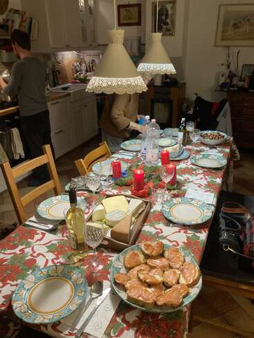 Uma mesa de jantar festiva está posta com pratos florais, velas, vinho, queijo e aperitivos de salmão. Duas pessoas estão preparando o jantar em uma cozinha quente e aconchegante.