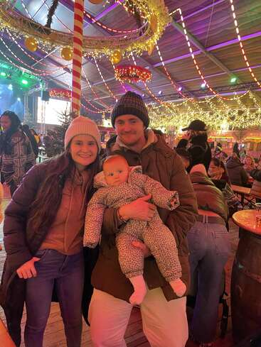 A smiling couple and their baby pose in a festive, warmly lit indoor event space decorated with Christmas lights, garlands, and people enjoying the cheerful, holiday atmosphere.