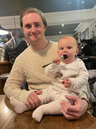 A smiling man holds a baby on his lap, inside a cozy restaurant. The baby, dressed in white, playfully chews on a spoon. Warm atmosphere.