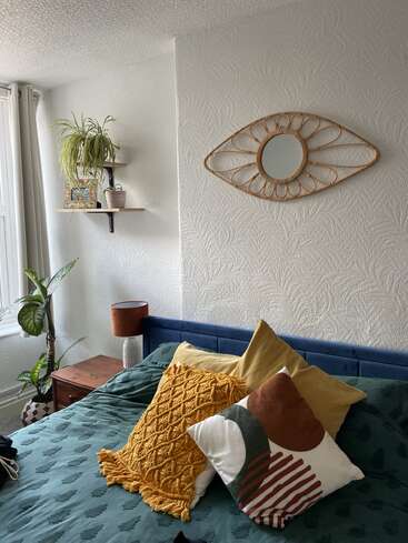 This cozy bedroom features a green bedspread, colorful pillows, houseplants, a wooden nightstand, and a unique eye-shaped wall mirror. Natural light and textures enhance coziness.