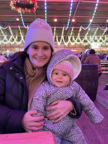 A woman and baby, warmly dressed in winter clothing, sit at a festive indoor venue with colorful lights and hanging decorations, smiling happily for the camera.