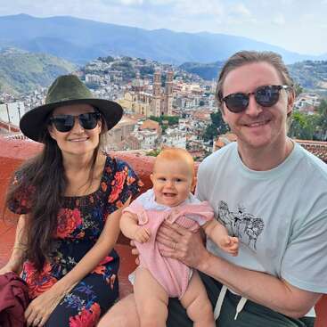 A smiling family of three, including a baby, posing for a photo with a scenic mountain town and church in the background on a sunny day.