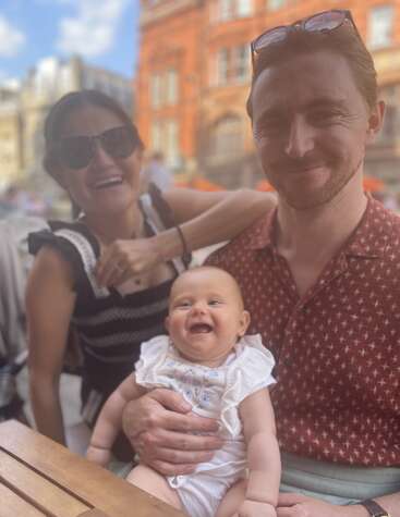 Un hombre y una mujer sonrientes están sentados al aire libre en una mesa de madera con un bebé feliz en el regazo del hombre, disfrutando de un día soleado en la ciudad.