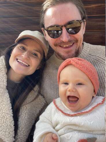 Una alegre familia de tres posa para una selfie al aire libre. La madre, el padre y el bebé sonríen, irradiando felicidad y calidez con ropa cómoda.