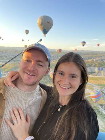 Una pareja sonriente posa para una selfie con coloridos globos aerostáticos flotando en el azul del cielo matutino sobre un paisaje pintoresco, creando una atmósfera alegre y aventurera.