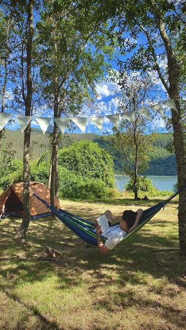 Une personne se détend dans un hamac, lisant un livre sous les arbres, près d'une tente et d'un lac. Des banderoles sont suspendues au-dessus d'elle et la lumière du soleil filtre à travers le feuillage.