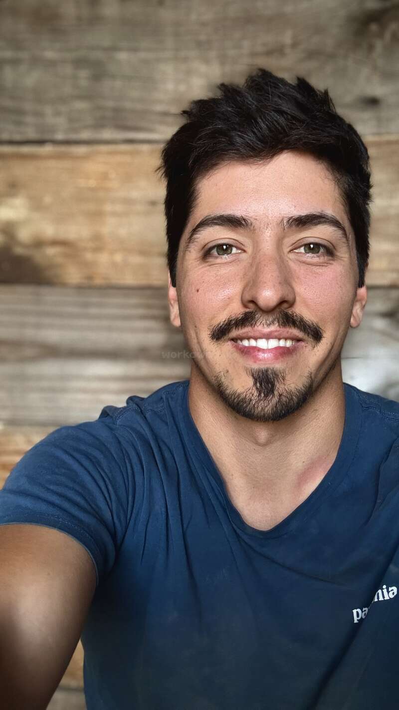 Un jeune homme souriant prend un selfie devant un fond en bois. Il a les cheveux courts et foncés, une barbe et porte une chemise bleue.