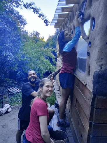 Trois personnes souriantes construisent ensemble un mur en terre naturelle en plein air. Entourées de verdure, elles semblent joyeuses et collaborer, travaillant avec de la boue et des matériaux durables.
