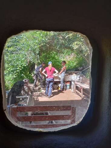 Quatre personnes travaillent en plein air dans un jardin, vu à travers une fenêtre. Elles semblent collaborer à un projet impliquant de la terre ou des matériaux de construction.