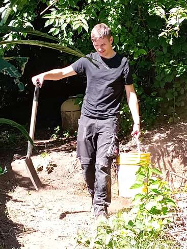 Un jeune homme vêtu de noir se tient à l'extérieur, une pelle et un seau jaune à la main. Il semble travailler dans un jardin entouré de feuillage vert.