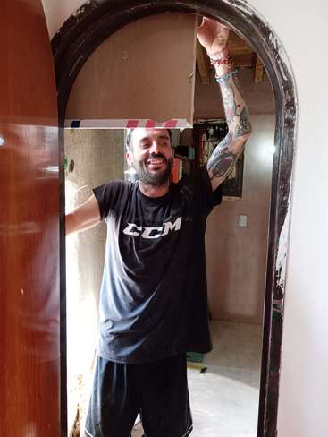 Un homme souriant et tatoué se tient dans l'embrasure d'une porte surmontée d'une découpe, partiellement bloquée par du ruban adhésif. Il porte un t-shirt CCM noir et un short, et s'amuse.