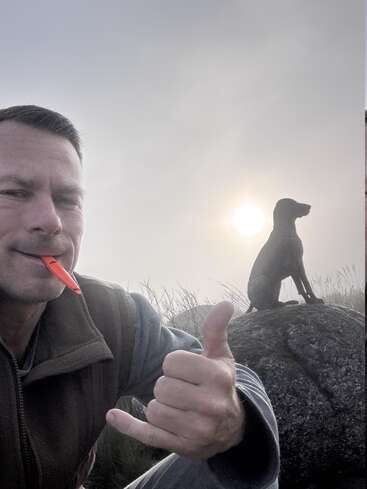 Um homem sorri para a câmera, fazendo o sinal de shaka com um apito na boca. Atrás dele, um cachorro está sentado em uma pedra ao nascer do sol.