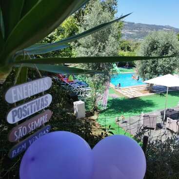 Uma cena ensolarada à beira da piscina com pessoas relaxando, árvores verdes, balões roxos e uma placa em português dando as boas-vindas às energias positivas. Montanhas e céu azul ao fundo.