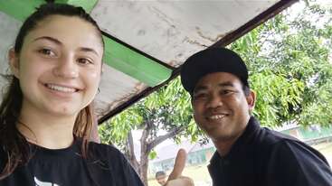 Une jeune femme et un homme sourient pour un selfie sous un abri. L'homme lève le pouce. On aperçoit des arbres et des bâtiments en arrière-plan.