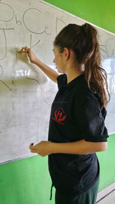 Une jeune femme écrit de grandes lettres sur un tableau blanc dans une salle de classe. Vêtue d'un t-shirt noir, elle se tient debout devant un mur vert et enseigne les bases de l'alphabet.