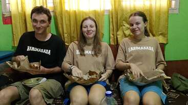 Trois jeunes sont assis sur un canapé, souriants, en train de manger dans leurs assiettes. Ils portent des t-shirts « HARIMASADA ». En arrière-plan, on aperçoit des rideaux jaunes et des murs verts.