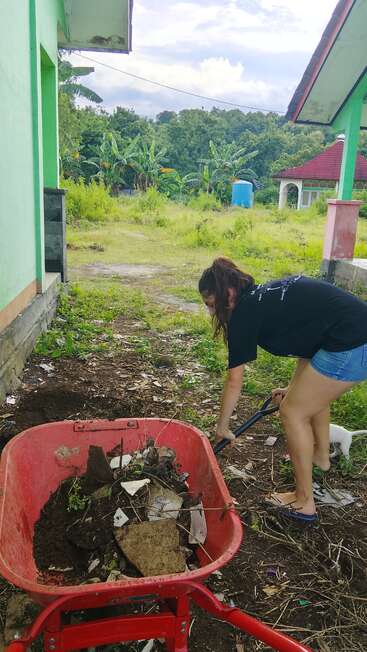 Une femme vêtue d'un t-shirt noir et d'un short nettoie un jardin ; elle utilise une pelle pour ramasser les déchets et les mettre dans une brouette rouge. Elle est entourée de verdure.