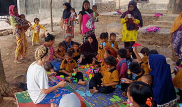 L'image représente un groupe d'enfants et d'adultes assis sur un tapis coloré, jouant avec des blocs de construction, entourés de quelques femmes et enfants à l'arrière-plan.