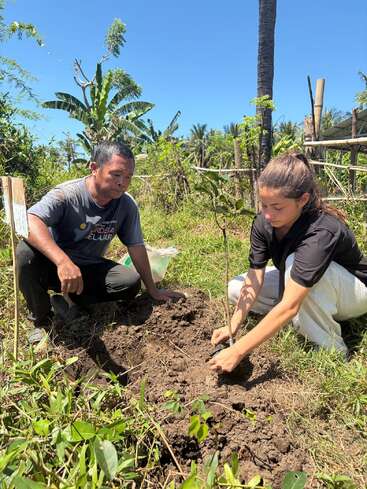 Un homme et une femme plantent ensemble un jeune arbre dans un champ verdoyant et ensoleillé, entourés de plantes, d'arbres et d'une clôture en bois.