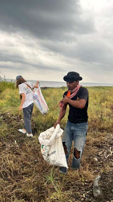 Deux personnes nettoient une pelouse au bord de l'eau ; sous un ciel nuageux, elles ramassent les déchets dans de grands sacs, contribuant ainsi à la protection de l'environnement et à la vie associative.