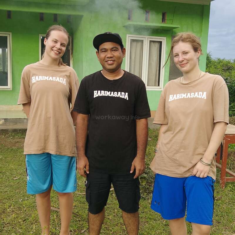 Trois personnes souriantes se tiennent devant un bâtiment vert, vêtues de t-shirts portant l'inscription « HARIMASADA ». Les deux femmes portent des t-shirts beiges, tandis que l'homme au milieu porte un t-shirt noir.