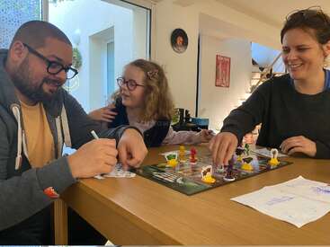 Tres personas, entre ellas un niño, juegan alegremente a un juego de mesa en una mesa situada en un salón acogedor y bien iluminado, compartiendo risas y sonrisas.