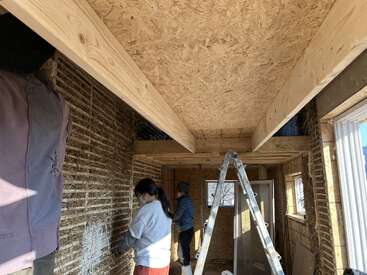 Three people work inside a partially constructed house, applying material to straw walls. A ladder stands in the center. Exposed beams and wood ceiling are visible throughout.
