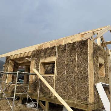 Una casa parcialmente construida con estructura de madera y aislamiento de paja. Se ven las aberturas de las ventanas, hay andamios y la estructura del techo aún no está terminada. Cielo nublado.