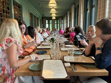 Un grand groupe de femmes est assis à une longue table de restaurant, engageant une conversation animée, entouré de menus, de verres et de la lumière naturelle provenant de hautes fenêtres.
