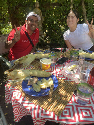Deux personnes sourient et font des signes de paix en dégustant un repas en plein air. Des épis de maïs, de l'eau et de la nourriture sont disposés sur une nappe à motifs, à l'ombre d'un feuillage.
