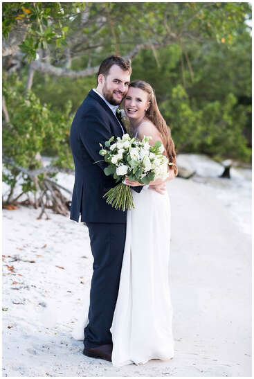 Des jeunes mariés joyeux s'embrassent sur une plage de sable, entourés de verdure. La mariée tient un grand bouquet de fleurs blanches, et tous deux sourient joyeusement.