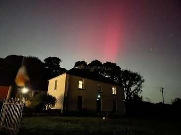 Une maison confortable se trouve sous un ciel nocturne, illuminée par une aurore rose saisissante, avec des arbres environnants et une atmosphère lumineuse créant une scène paisible.