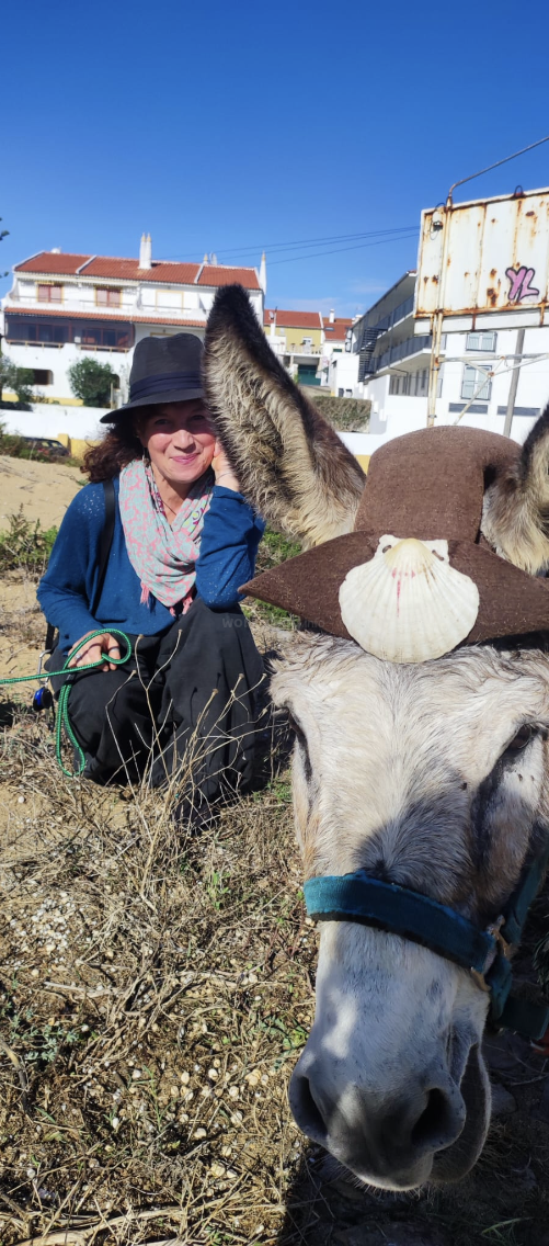 La imagen muestra a una mujer arrodillada junto a un burro adornado con un sombrero marrón con una concha marina, sobre un fondo de edificios y un cielo azul despejado.