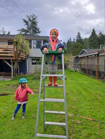 Deux enfants jouent dehors dans un jardin herbeux. L'un d'eux, vêtu d'une veste à capuche, est debout en haut d'une échelle métallique ; l'autre, qui porte un casque, l'observe d'en bas.