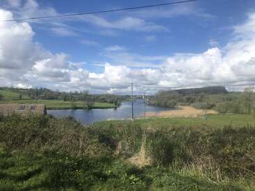 Das Bild zeigt eine ruhige Landschaft mit einem Fluss, üppigem Grün und einem blauen Himmel mit weißen Wolken, die ein Gefühl von Ruhe und natürlicher Schönheit hervorrufen.