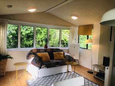Das Bild zeigt ein gemütliches Wohnzimmer mit einer weißen Couch, gelben Akzenten und einem großen Fenster, das einen ruhigen Blick auf die üppigen grünen Bäume draußen bietet.