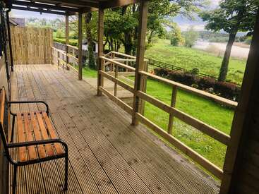 Das Bild zeigt eine ruhige Holzterrasse mit einer Bank, die einen Blick auf ein üppiges grünes Feld und Bäume bietet und ein Gefühl von Ruhe und natürlicher Schönheit ausstrahlt.