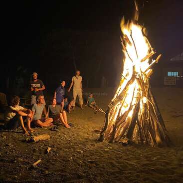 Eine Gruppe von Menschen sitzt nachts um ein großes Lagerfeuer am Strand und genießt die Wärme und das Licht der Flammen. Das Feuer ist groß und hell.