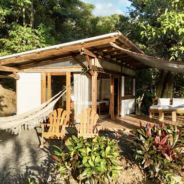 A cozy wooden cabin sits in lush greenery, featuring a hammock, two wooden chairs, outdoor seating, large windows, and a peaceful, inviting natural atmosphere.