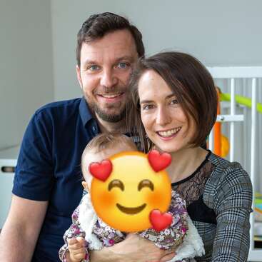 Das Bild zeigt eine dreiköpfige Familie, die für ein Foto posiert. Die Mutter hält ein Baby und der Vater steht lächelnd hinter ihnen.