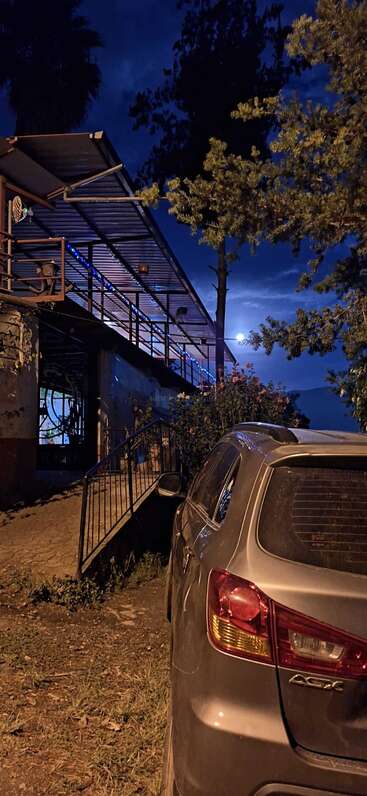 Ein metallfarbenes Auto der Marke ASX steht neben einem Haus mit blauen Lichtern. Bäume, Blumen und ein heller Vollmond machen die friedliche Nachtlandschaft hell.