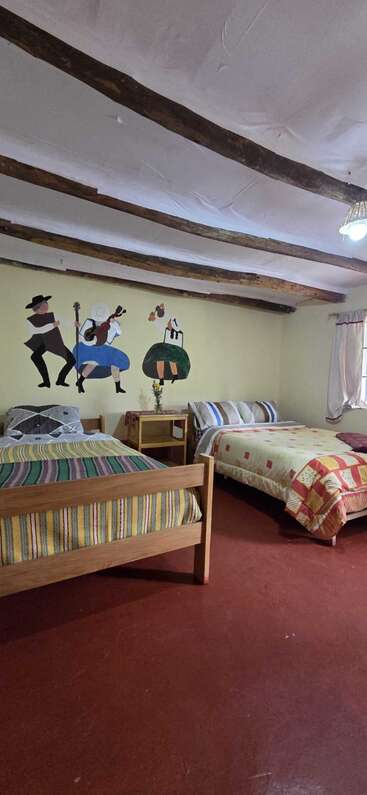 Dieses gemütliche Zimmer hat zwei Holzbetten mit bunten Decken, rustikale Deckenbalken, ein Wandbild mit Tänzern und Musikern und einen kleinen Tisch mit Blumen.