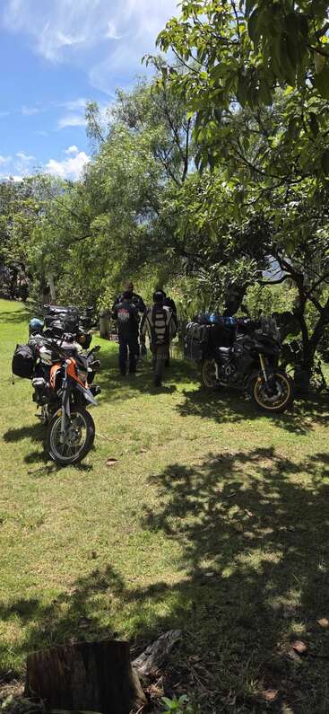 Drei Motorräder mit Ausrüstung stehen auf einer Wiese unter Bäumen. Zwei Leute in Motorradklamotten quatschen und genießen die sonnige, ruhige Atmosphäre draußen.