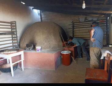 Dois caras trabalham numa padaria rústica, usando um forno tradicional de barro. Prateleiras de metal, tabuleiros de cozedura, uma mesa e recipientes cercam o espaço de trabalho. O ambiente é acolhedor e autêntico.