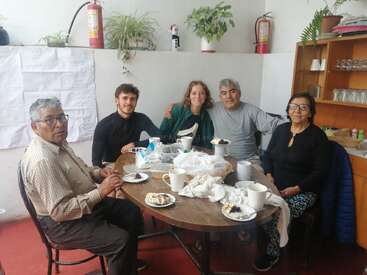 Cinco pessoas sentadas em torno de uma mesa de madeira, saboreando bolo e bebidas. O ambiente parece aconchegante, com plantas de interior e prateleiras, sugerindo uma reunião informal e amigável.