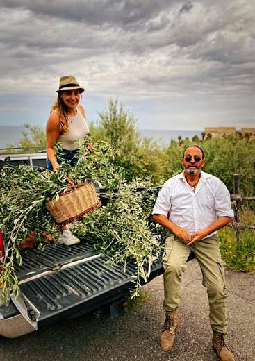 A woman stands on the back of a truck, holding a basket of olive branches, with a man sitting beside her in a scenic outdoor setting.