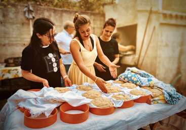 The image shows a group of people gathered around a table, engaged in a baking activity, with a large bowl of dough and a baking sheet in the background, likely for a community event or gathering.