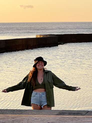The image depicts a woman standing on a stone pier, arms outstretched, with her back to the ocean, wearing a hat, jacket, and shorts, in front of a calm sea at sunset.